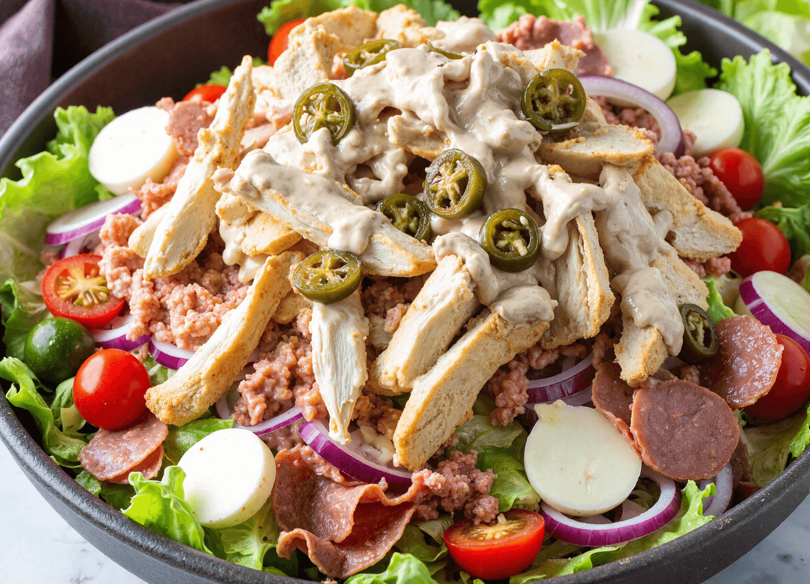 Close-up of Italian grinder salad with sliced turkey, salami, hearts of palm, cherry tomatoes, and creamy dressing in a white bowl on a wooden surface.