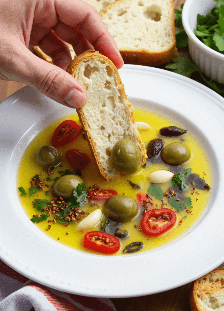 Hand dipping crusty bread into a loaded bread dipping oil with green olives, garlic, herbs, and tomatoes in a white bowl.