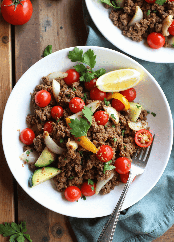 Overhead shot of Mediterranean ground beef stir fry served in a white bowl with cherry tomatoes, sautéed onions, zucchini, yellow bell peppers, and a lemon wedge, highlighting a quick and healthy weeknight dinner.