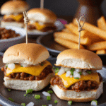 Close-up of two mini chili cheese dog sliders topped with cheddar cheese, onions, and jalapeños on a black plate with fries in the background.