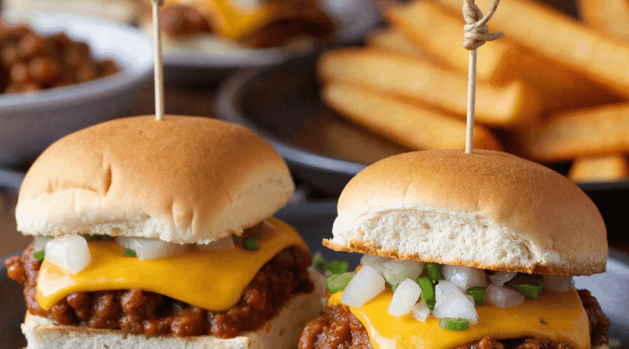 Close-up of two mini chili cheese dog sliders topped with cheddar cheese, onions, and jalapeños on a black plate with fries in the background.