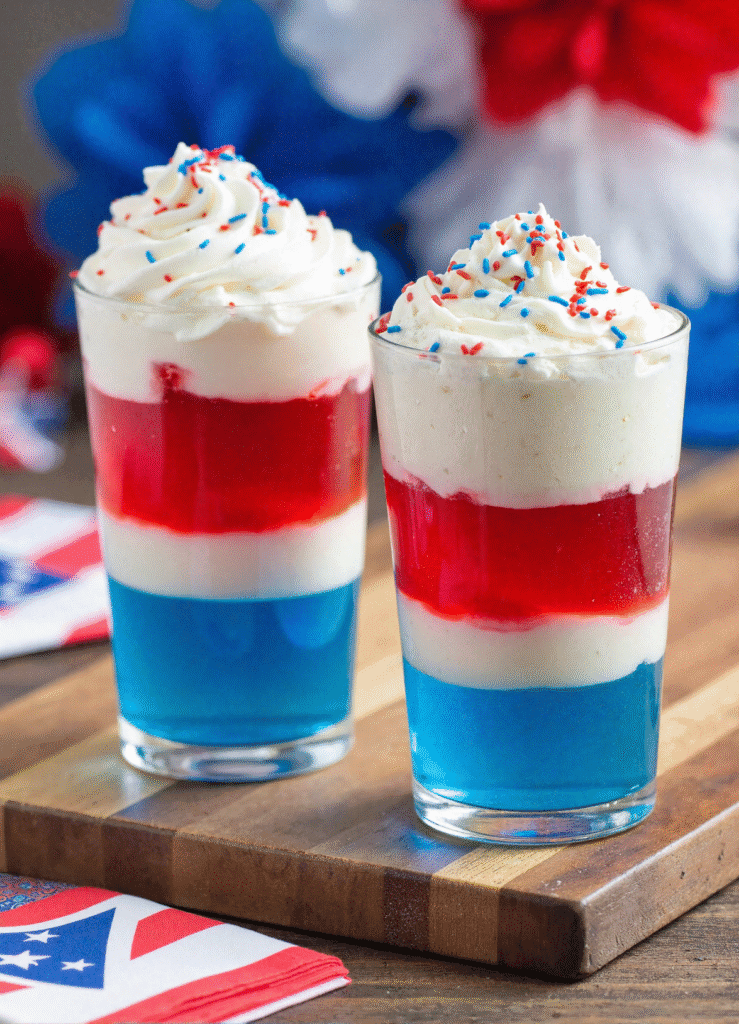 Layered red, white, and blue jello dessert cups topped with whipped cream and star sprinkles, perfect for Fourth of July celebrations.