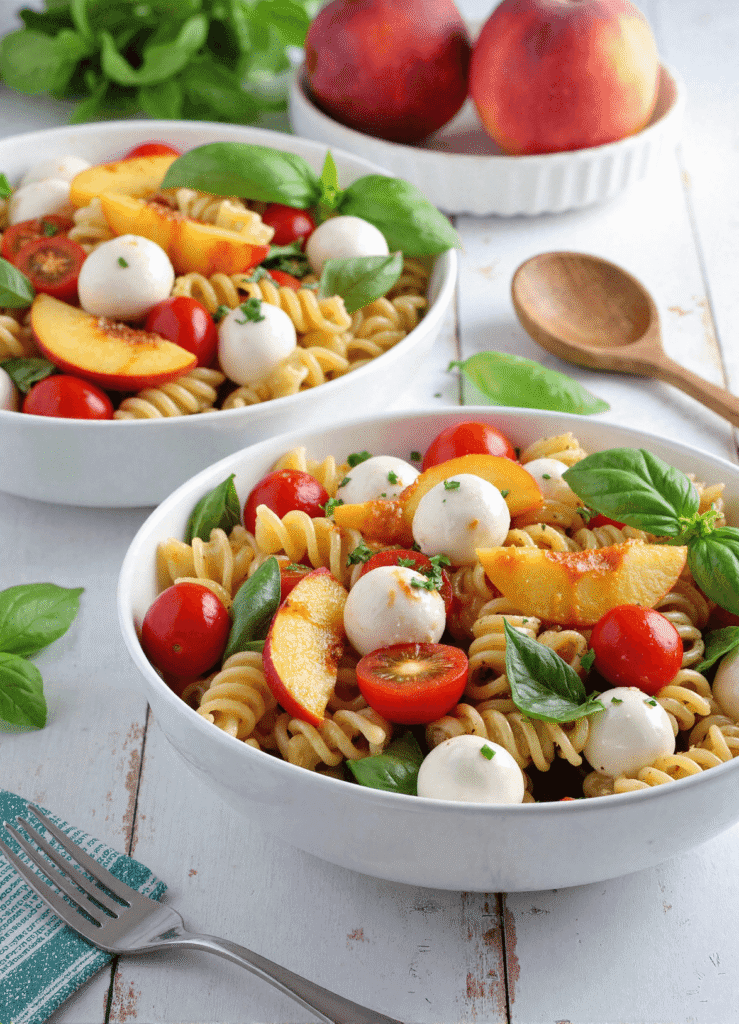 Bowl of peach Caprese pasta salad with fresh basil, cherry tomatoes, mozzarella balls, and rotini pasta on a white rustic table.