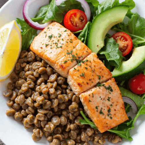 Healthy salmon and lentil salad bowl with leafy greens, cherry tomatoes, avocado slices, lemon wedge, and seasoned grilled salmon.
