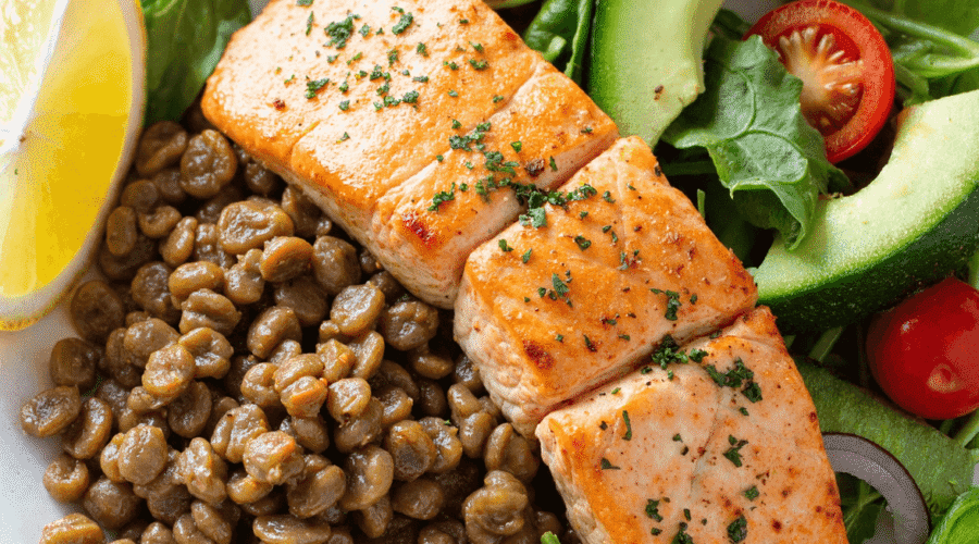 Healthy salmon and lentil salad bowl with leafy greens, cherry tomatoes, avocado slices, lemon wedge, and seasoned grilled salmon.