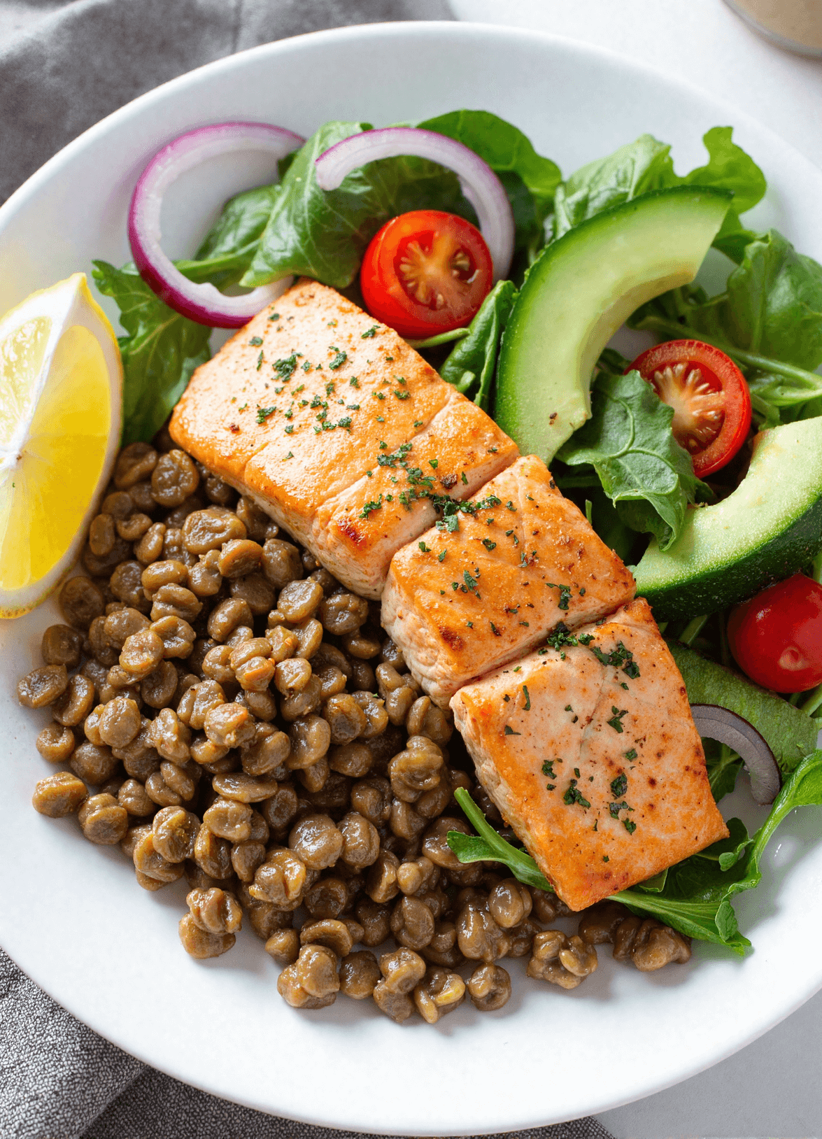 Healthy salmon and lentil salad bowl with leafy greens, cherry tomatoes, avocado slices, lemon wedge, and seasoned grilled salmon.