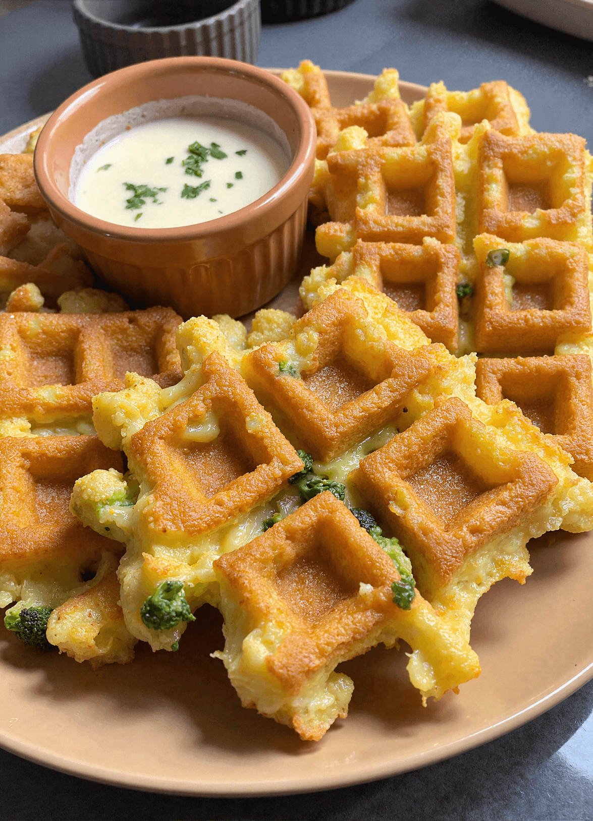 Crispy broccoli and cheddar waffles arranged around a ramekin of creamy dipping sauce on a tan plate.