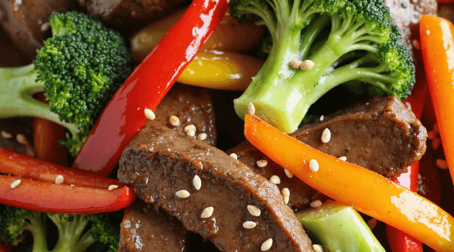 Sliced beef stir-fry with broccoli, red and yellow peppers, served over white rice and topped with sesame seeds.