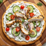 Whole flatbread pizza topped with tzatziki sauce, grilled chicken, cucumbers, tomatoes, red onion, and fresh herbs.
