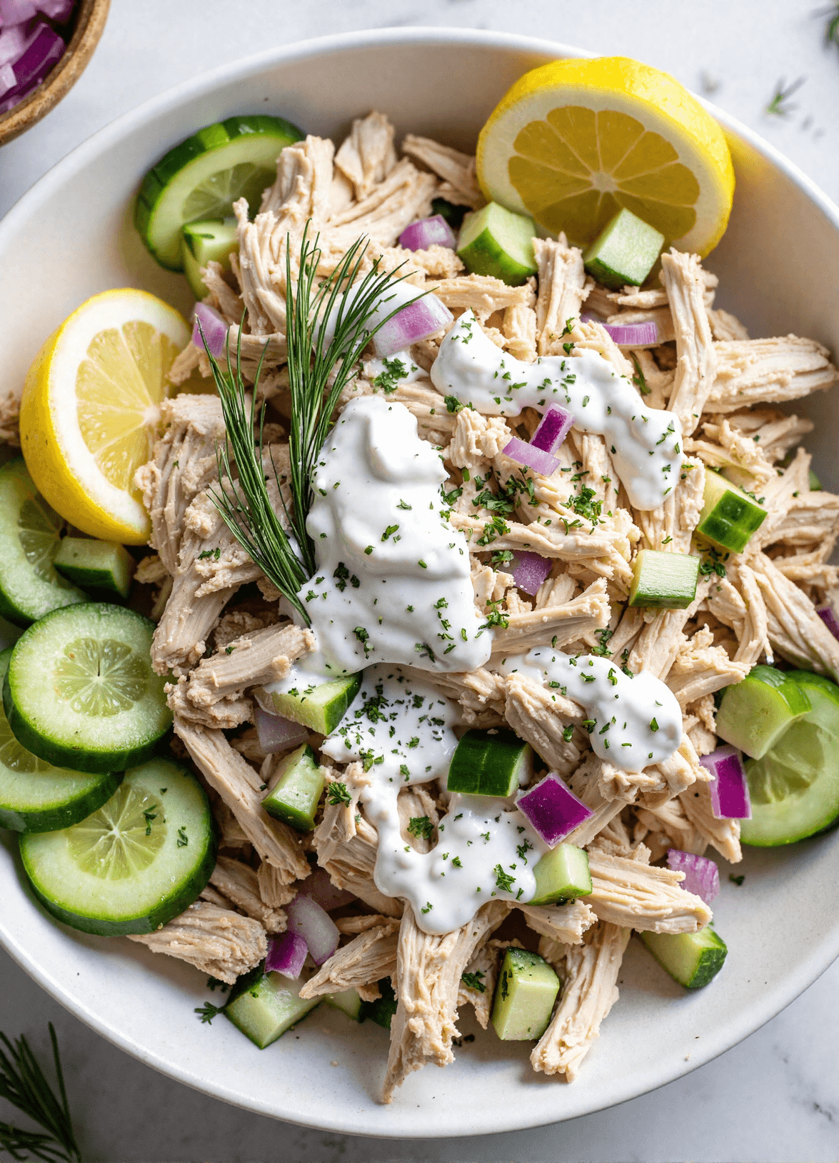 Bowl of creamy tzatziki chicken salad with cucumbers, red onion, dill, and lemon slices.