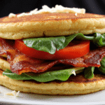 Layered BLT protein pancake sandwich filled with bacon, cheddar cheese, spinach, and tomatoes on a white plate.