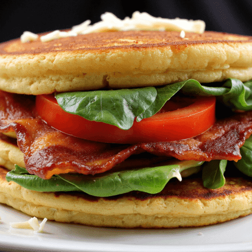 Layered BLT protein pancake sandwich filled with bacon, cheddar cheese, spinach, and tomatoes on a white plate.