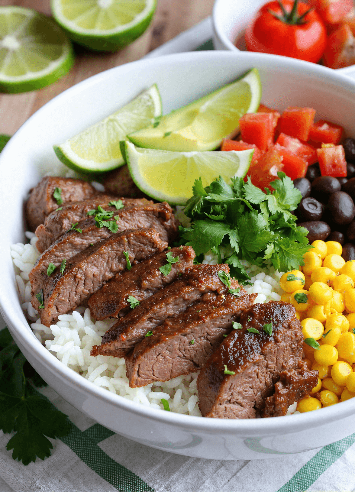 Cilantro lime steak bowl featuring sliced steak over rice with lime wedges, corn, black beans, diced tomato, and fresh cilantro for an easy Tex-Mex meal prep dish.