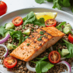 Grilled salmon atop a fresh salad of lentils, avocado, red onion, cherry tomatoes, and greens with lemon garnish on a white ceramic plate.
