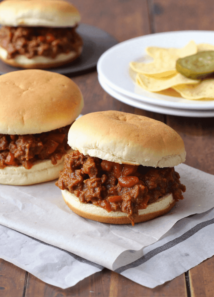 Three sloppy joe sandwiches on a rustic table with a side plate of potato chips and pickles in the background.