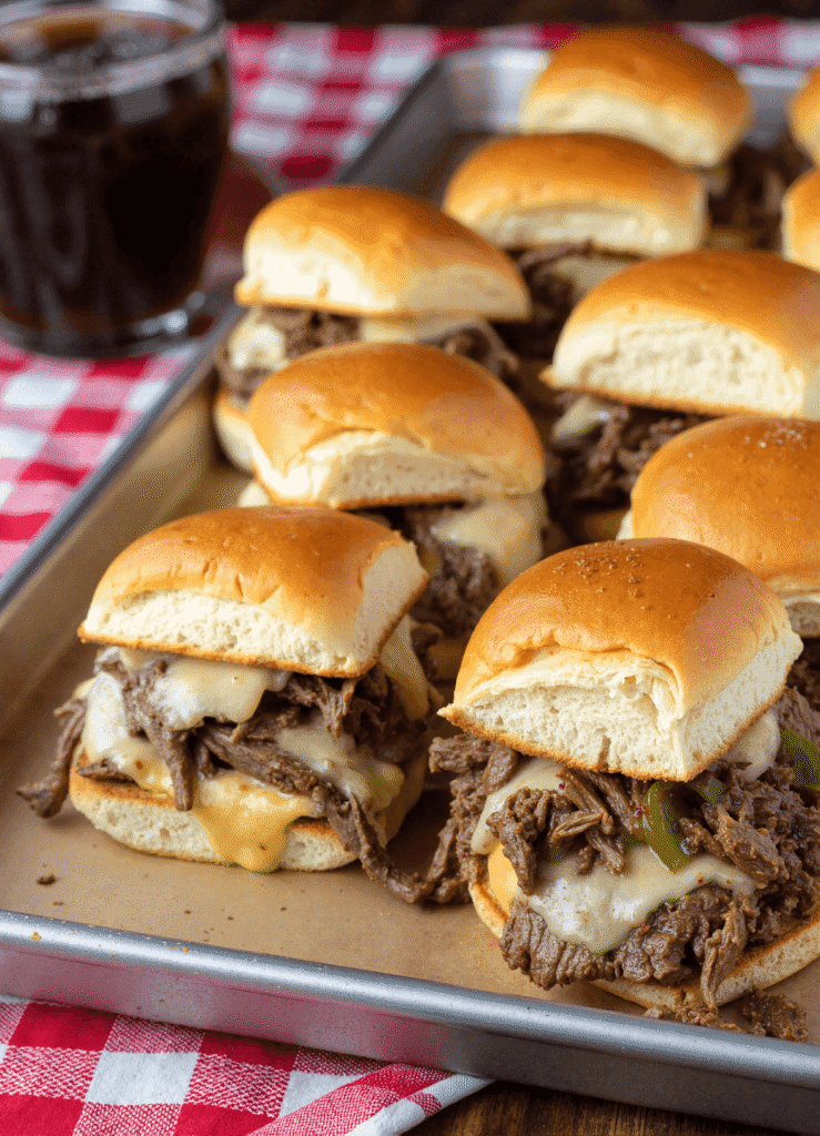 Philly cheesesteak sliders on a baking sheet, filled with melted provolone, shaved beef, and bell peppers on golden slider buns