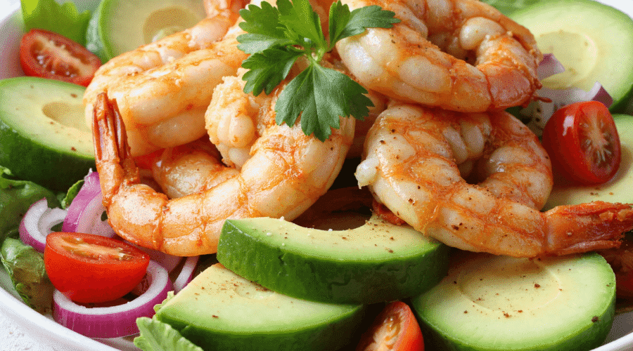 Close-up of shrimp and avocado salad with fresh greens, cherry tomatoes, and red onions on a white plate.