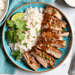 Sliced ginger lime pork loin drizzled with sticky glaze, plated next to fluffy coconut rice with fresh herbs and a lime wedge.