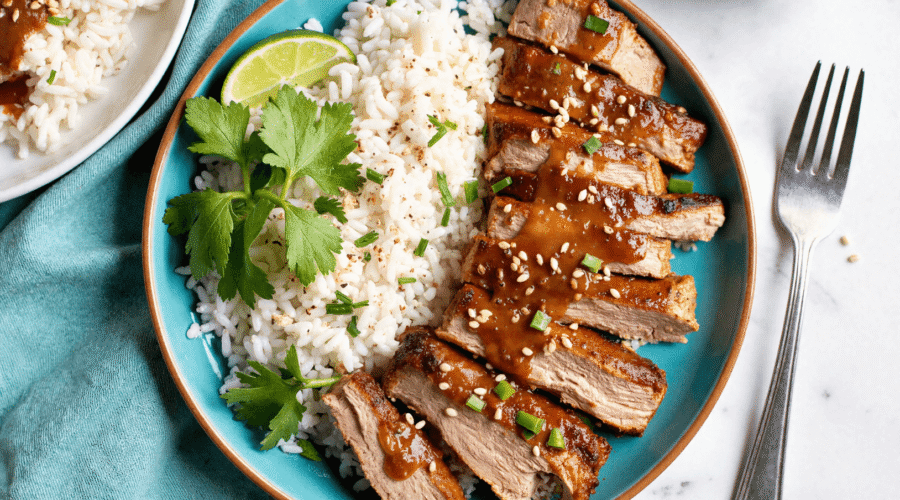 Sliced ginger lime pork loin drizzled with sticky glaze, plated next to fluffy coconut rice with fresh herbs and a lime wedge.