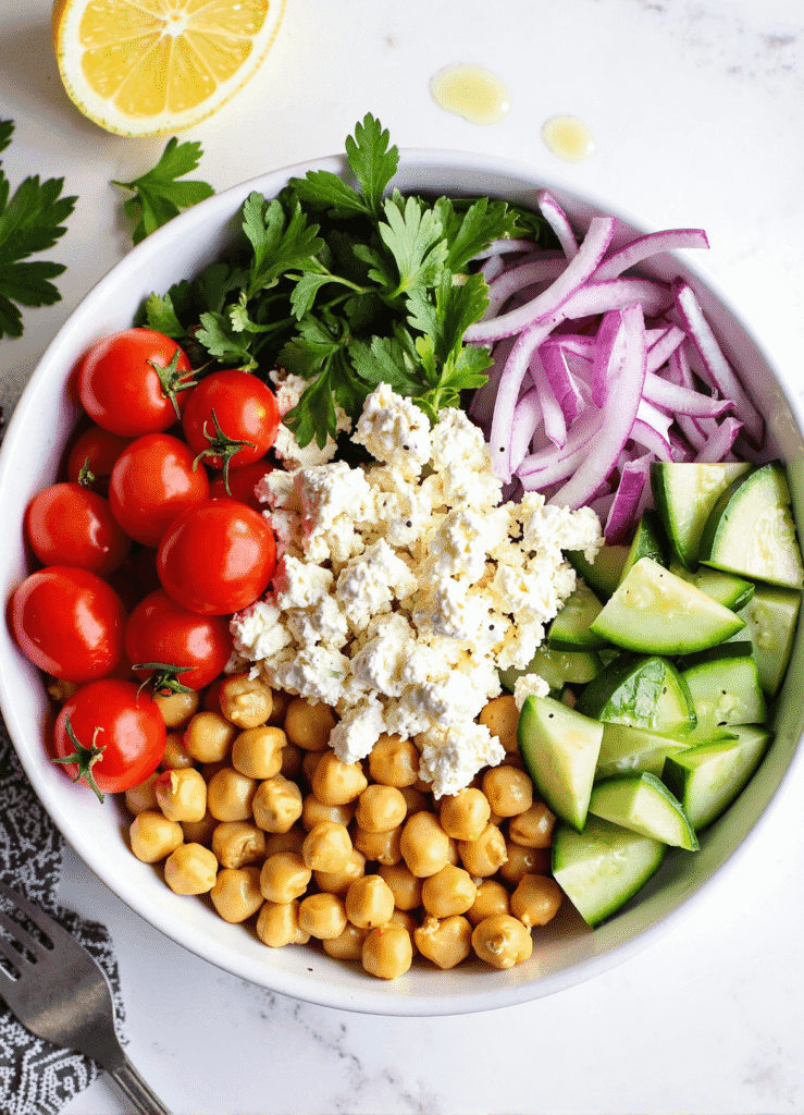Overhead view of a fresh chickpea cucumber feta salad with cherry tomatoes, red onion, parsley, and lemon in a white bowl.