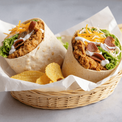 Two halved chicken bacon ranch wraps with crispy chicken, smoky bacon, fresh lettuce, shredded cheese, and creamy ranch dressing in a woven basket with potato chips.