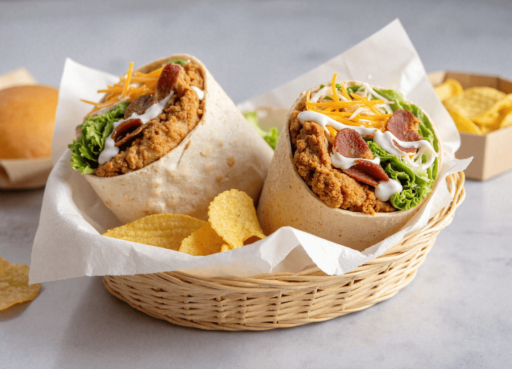 Two halved chicken bacon ranch wraps with crispy chicken, smoky bacon, fresh lettuce, shredded cheese, and creamy ranch dressing in a woven basket with potato chips.