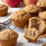 Apple cinnamon muffins on a cooling rack, with one muffin cut open to reveal moist interior and fruit pieces.