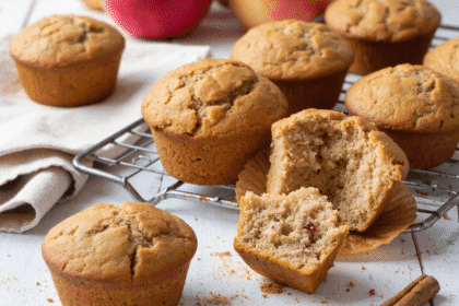 Apple cinnamon muffins on a cooling rack, with one muffin cut open to reveal moist interior and fruit pieces.