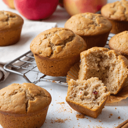 Apple cinnamon muffins on a cooling rack, with one muffin cut open to reveal moist interior and fruit pieces.