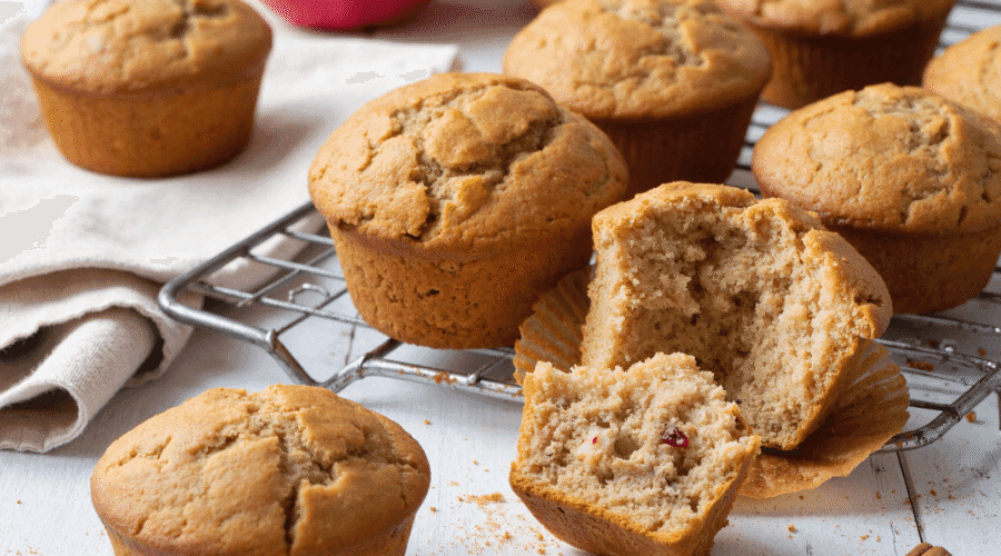 Apple cinnamon muffins on a cooling rack, with one muffin cut open to reveal moist interior and fruit pieces.