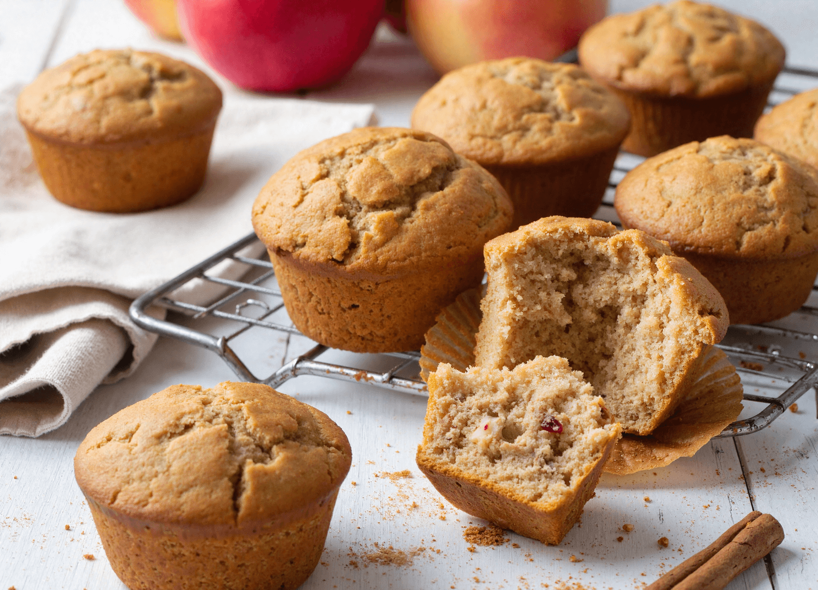 Apple cinnamon muffins on a cooling rack, with one muffin cut open to reveal moist interior and fruit pieces.