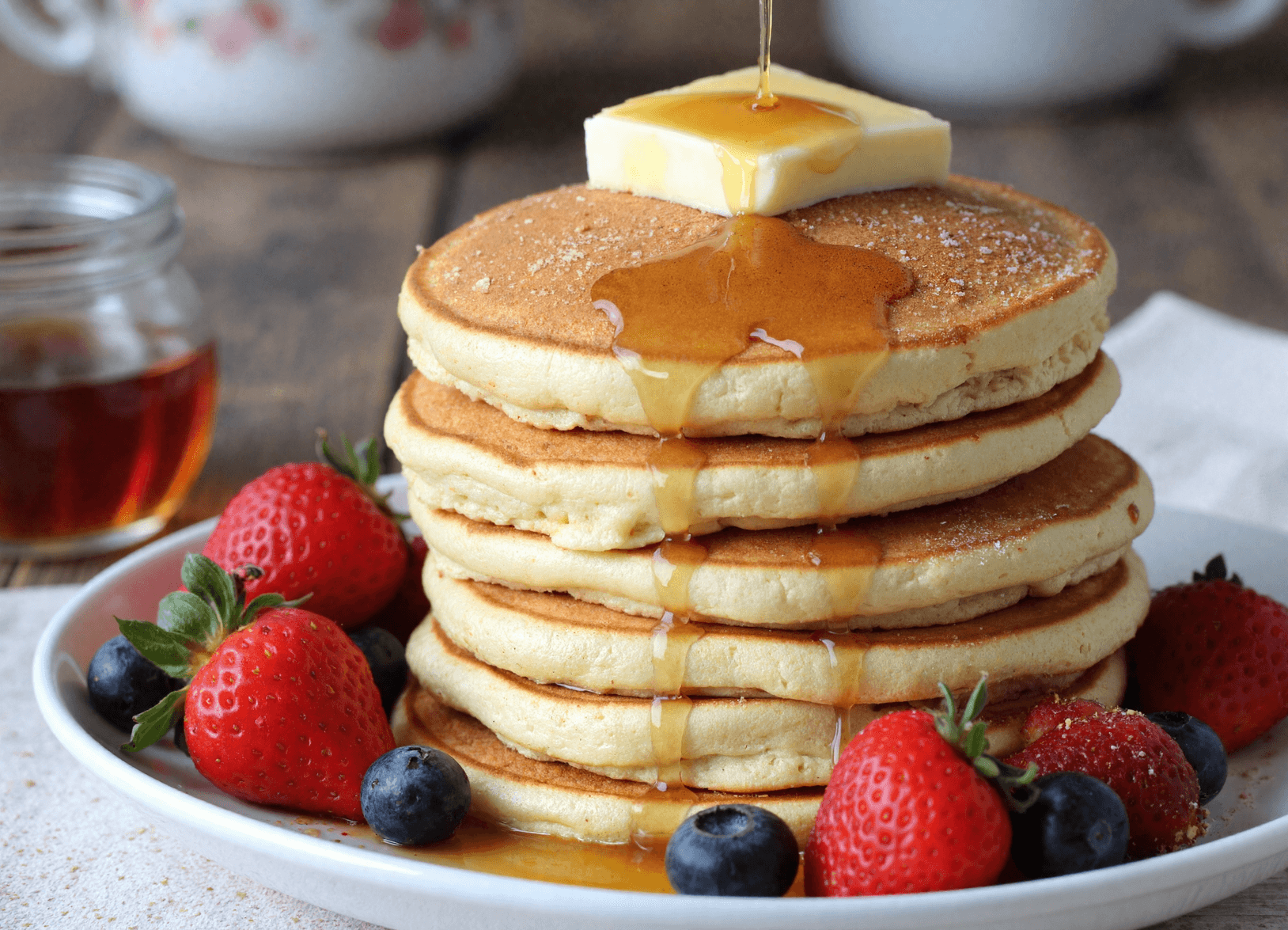 Stack of fluffy vanilla cinnamon pancakes with butter and maple syrup, surrounded by strawberries and blueberries.