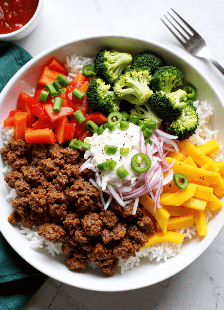 Ground beef rice bowl with broccoli, red peppers, onions, cheese, and green onions served in a white dish.