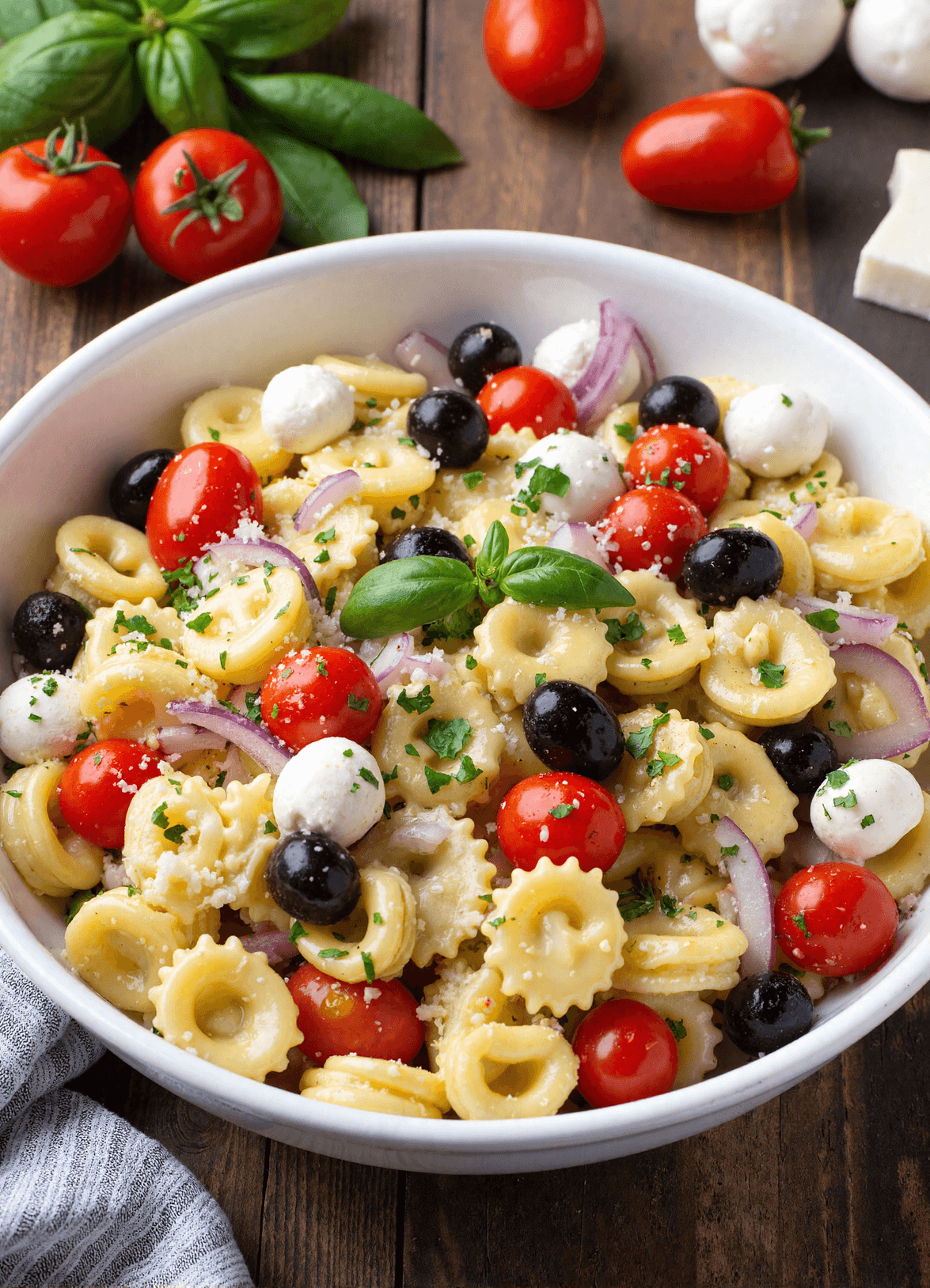 Italian pasta salad made with cheese tortellini, cherry tomatoes, black olives, red onions, and mini mozzarella balls in a white bowl.