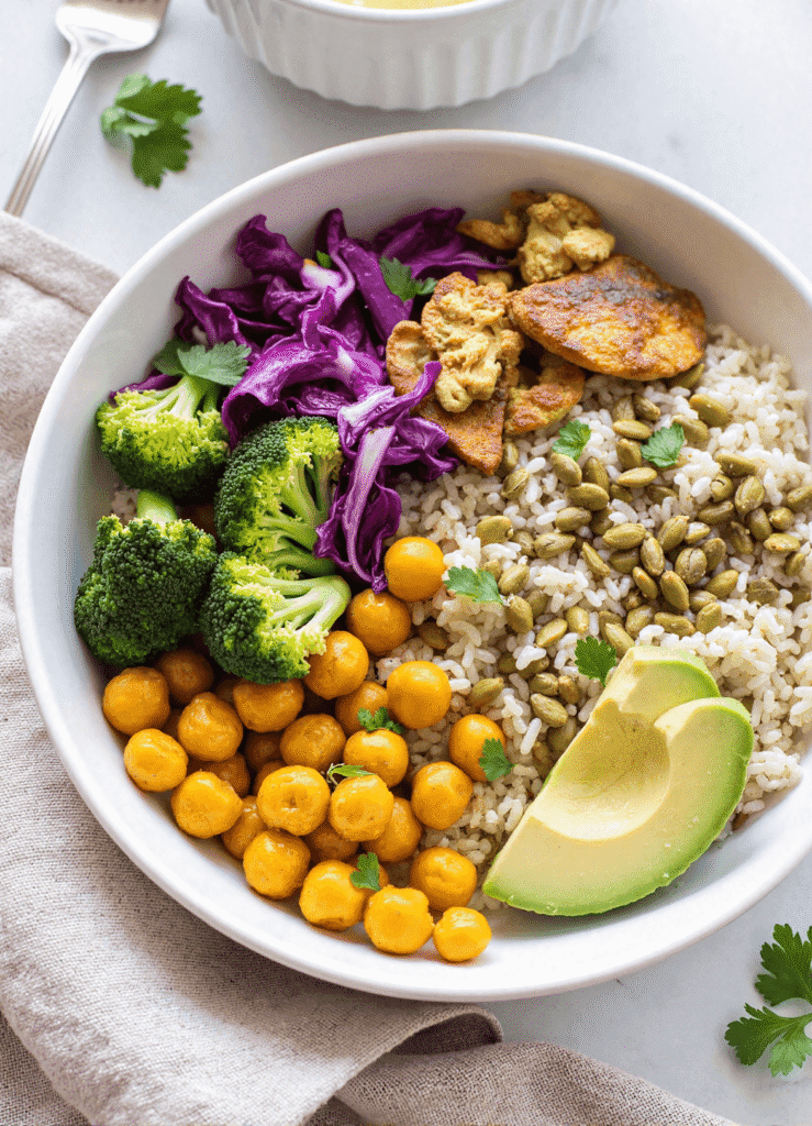 Overhead shot of a vibrant anti-inflammatory glow bowl with brown rice, roasted chickpeas, steamed broccoli, purple cabbage, turmeric cauliflower, pumpkin seeds, and avocado slices, arranged in a white bowl.