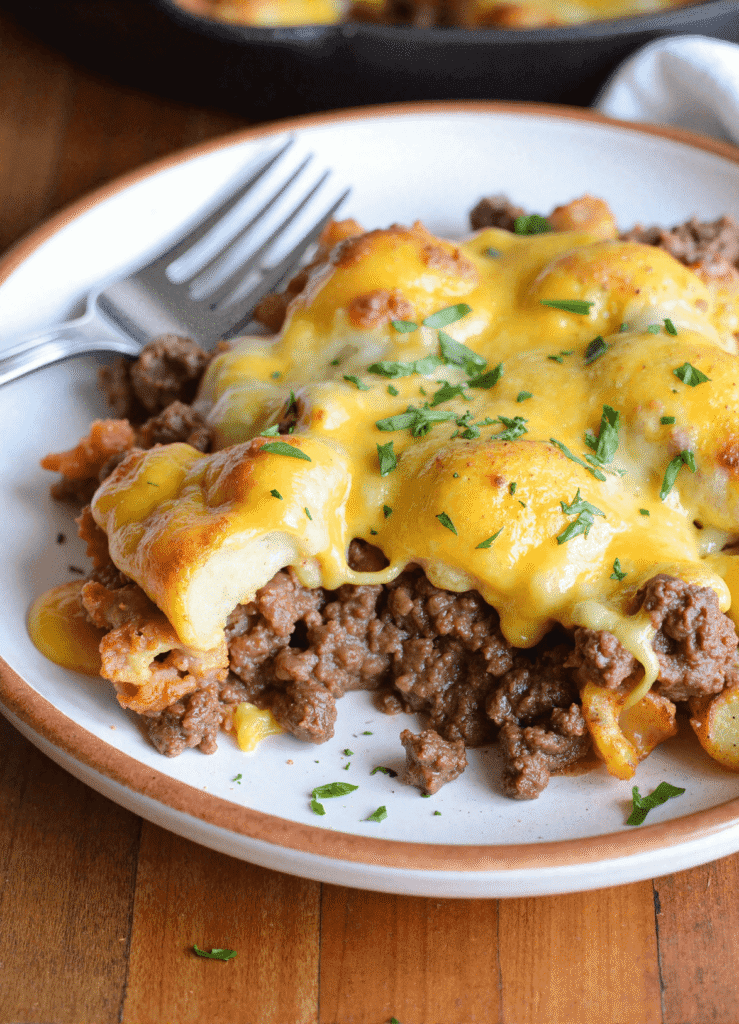 Close-up of a serving of cheesy ground beef and potato skillet on a white ceramic plate, topped with melted cheddar and fresh herbs.
