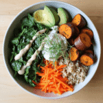 Bright overhead view of a nourishing bowl with roasted sweet potatoes, cooked quinoa, sautéed spinach, shredded carrots, avocado, creamy tahini dressing, and fresh herbs.