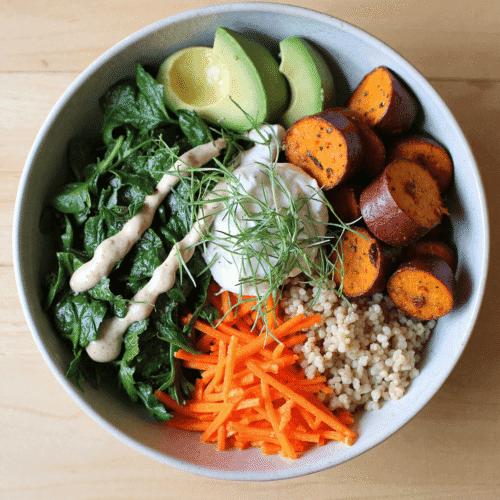 Bright overhead view of a nourishing bowl with roasted sweet potatoes, cooked quinoa, sautéed spinach, shredded carrots, avocado, creamy tahini dressing, and fresh herbs.