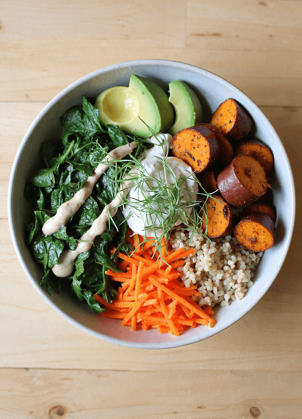 Bright overhead view of a nourishing bowl with roasted sweet potatoes, cooked quinoa, sautéed spinach, shredded carrots, avocado, creamy tahini dressing, and fresh herbs.