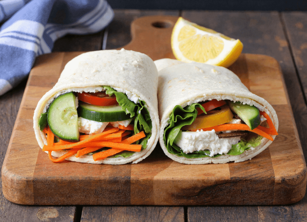 Two cottage cheese veggie wraps cut in half to show colorful layers of carrots, cucumbers, tomatoes, and creamy filling on a wooden board with a lemon wedge.