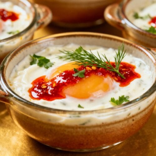 Baked Turkish egg pots in ceramic bowls, topped with poached eggs, chili butter, and herbs over garlic yogurt.