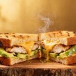 Hot grilled Chicken Avocado Melt Sandwich with golden bread, creamy avocado slices, juicy chicken breast, and melted cheese dripping onto a wooden board.