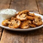 Golden fried mushroom slices with crunchy coating served with ranch dipping sauce on a white plate.