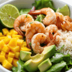 Fresh shrimp avocado mango bowl with rice, avocado slices, diced mango, and lime served over greens.