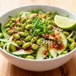 Spicy cucumber edamame salad with sesame seeds, chili flakes, soy dressing, and lime wedge in a white bowl.