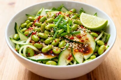 Spicy cucumber edamame salad with sesame seeds, chili flakes, soy dressing, and lime wedge in a white bowl.