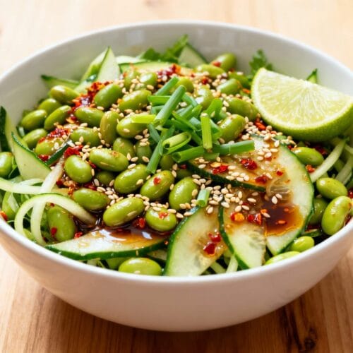 Spicy cucumber edamame salad with sesame seeds, chili flakes, soy dressing, and lime wedge in a white bowl.