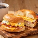 Breakfast bagel sandwich filled with scrambled eggs, melted cheese, crispy bacon, and creamy spread served with coffee and berries.