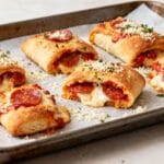 Golden-brown pepperoni pizza rolls filled with gooey mozzarella and lean protein, baked to perfection on a parchment-lined tray and topped with grated cheese and parsley.