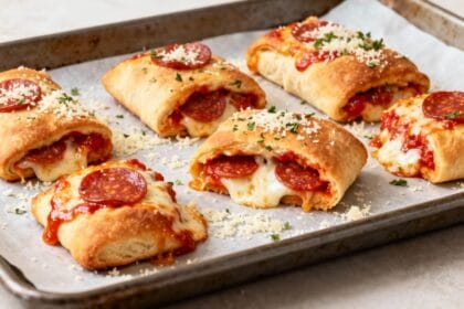 Golden-brown pepperoni pizza rolls filled with gooey mozzarella and lean protein, baked to perfection on a parchment-lined tray and topped with grated cheese and parsley.